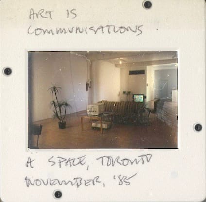 A sofa and chair positioned in front of a Teildon terminal and TV set in a large gallery space. Slide text reads: ART IS COMMUNICATIONS: A SPACE, TORONTO, NOVEMBER 1985.