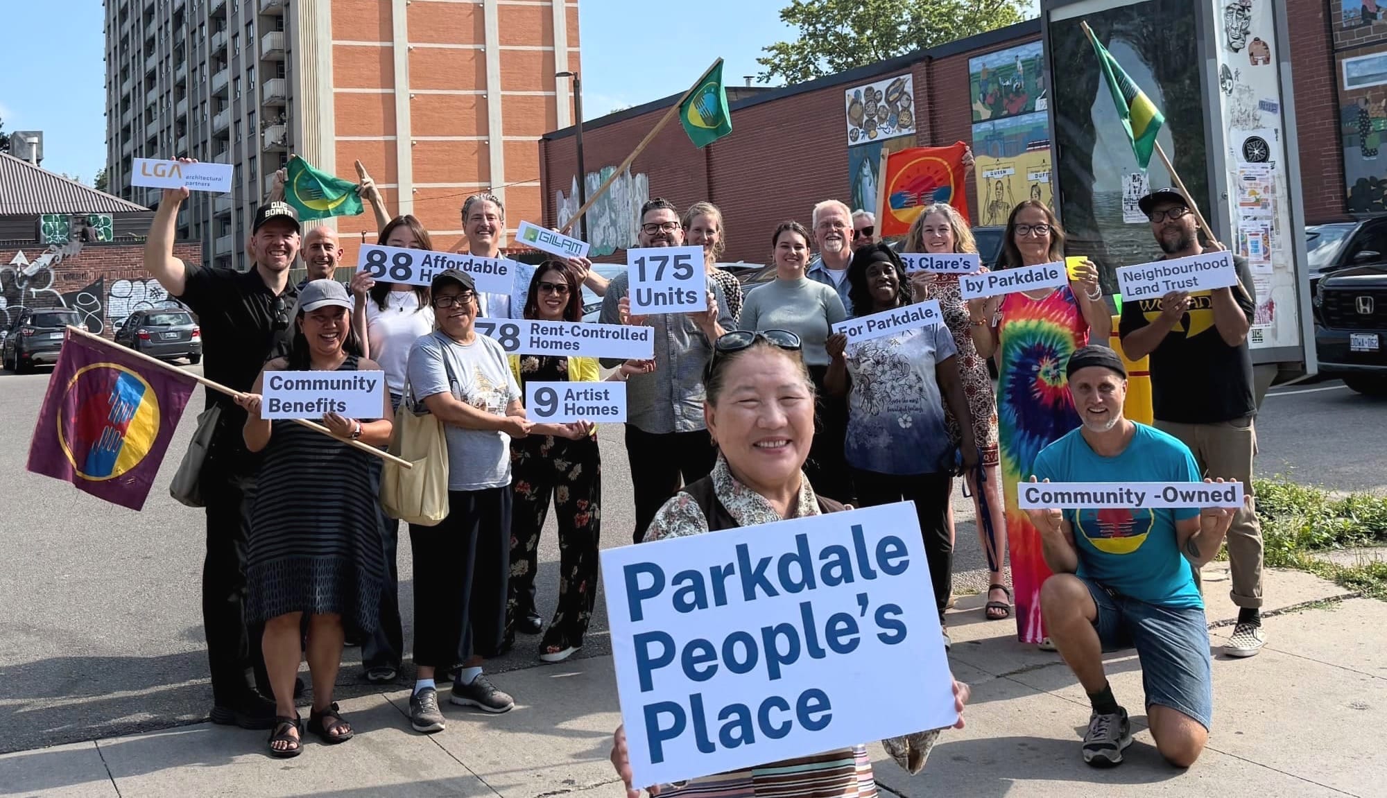 A group of smiling people in a parking lot, holding signs that read "Parkdale People's Place."