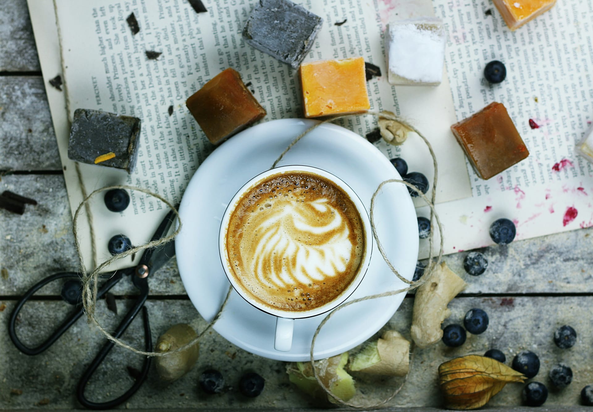 Latte on a white saucer with a backdrop of blueberries and art materials. Photo by Toa Heftiba via Unsplash.